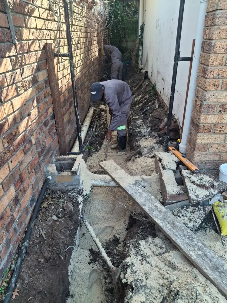 Hand-digging a trench along a house to expose and replace a buried waste line